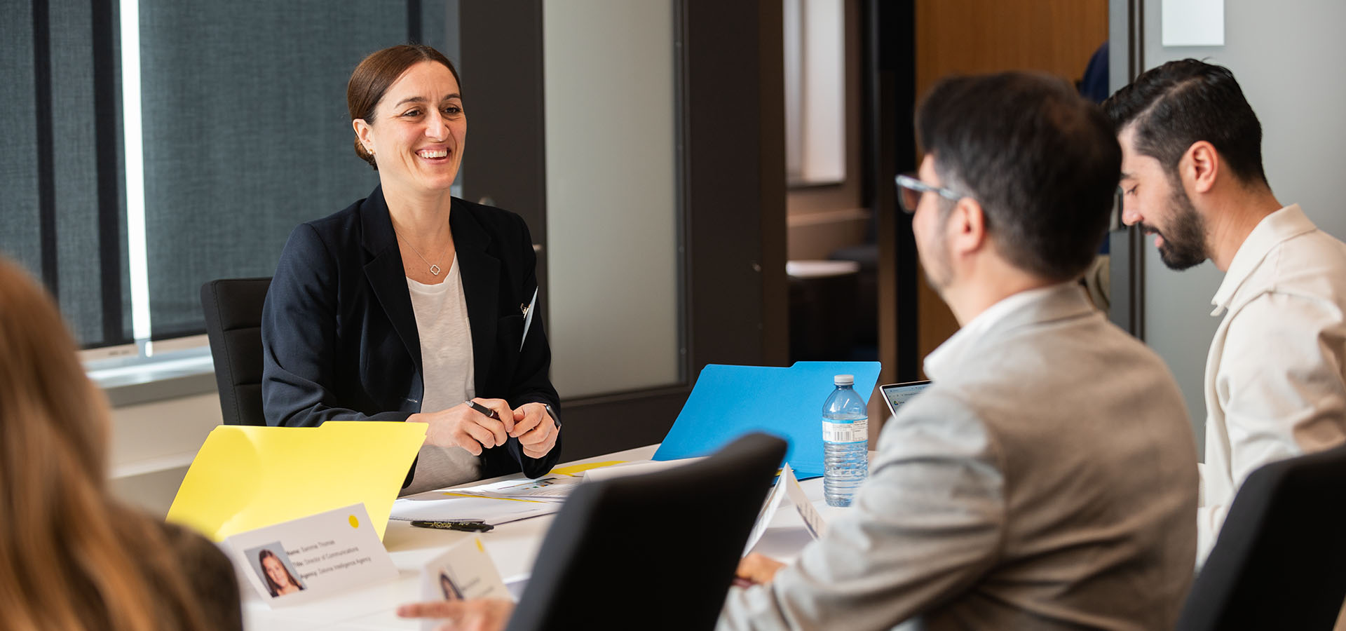 A group of colleagues are working together in a boardroom. A woman with brown hair smiles at her two male colleagues across the table.